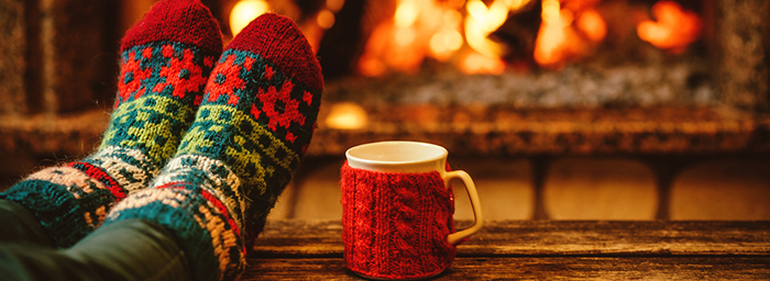 A close-cropped photo of someone's feet, covered in colourful knitted Christmas-themed socks, propped up on a coffee table in front of a fire in a fireplace. Also on the coffee table is a mug in a knitted cozy.