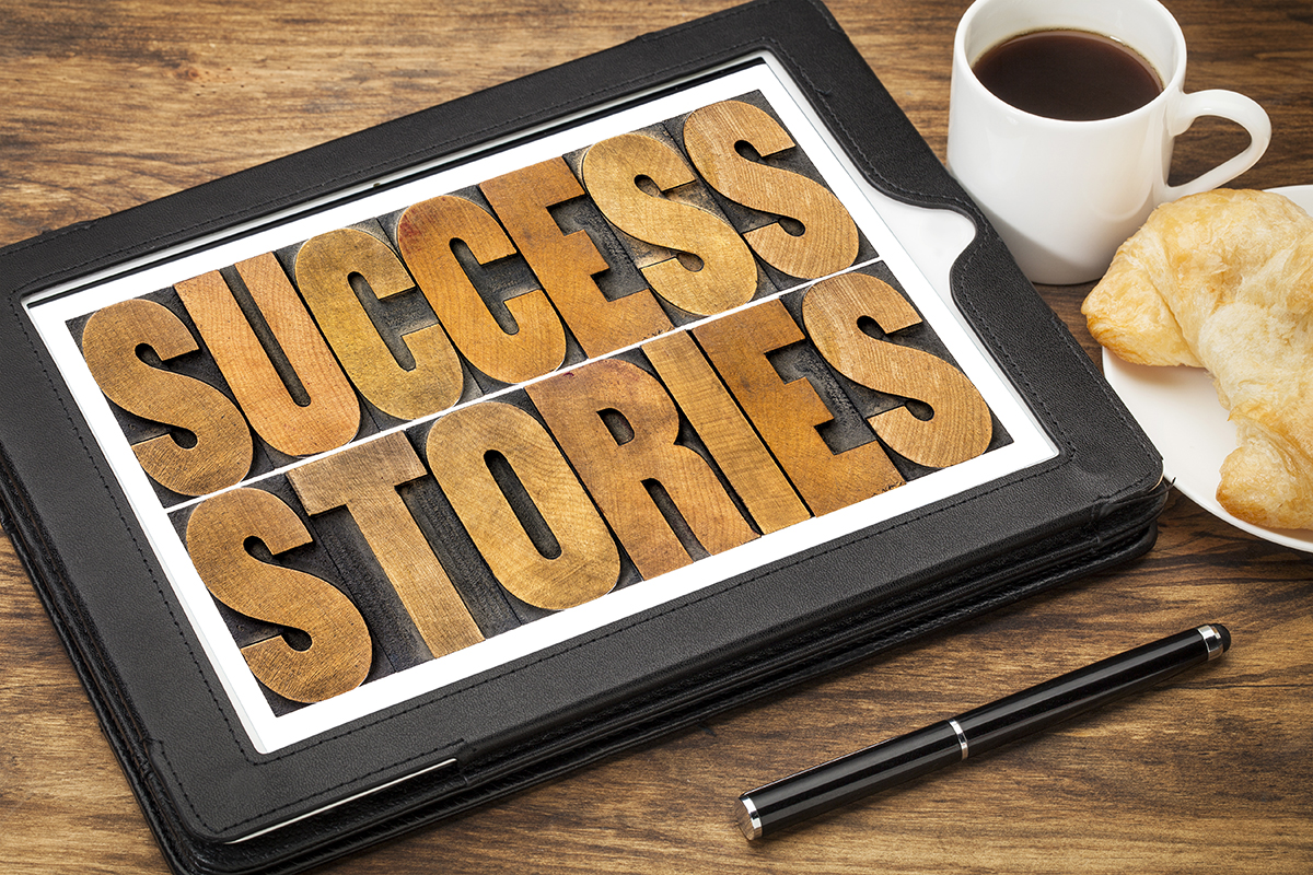 A tablet sits on a desk next to a cup of coffee and a croissant. Upon the tablet is a leather cover with the words Success Stories printed on it in lettering that looks like it is made out of stained wood.