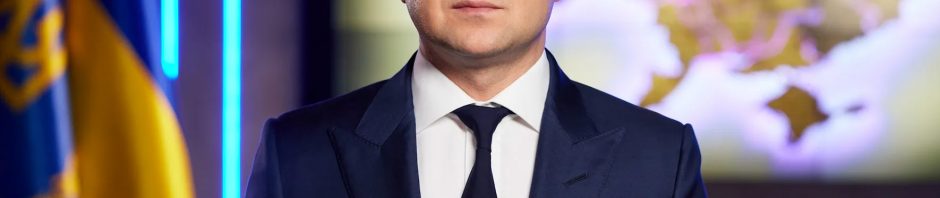 Ukraine President Volodymyr Zelenskyy faces the camera in a suit and tie. Ukraine flag in foreground. World map in background.