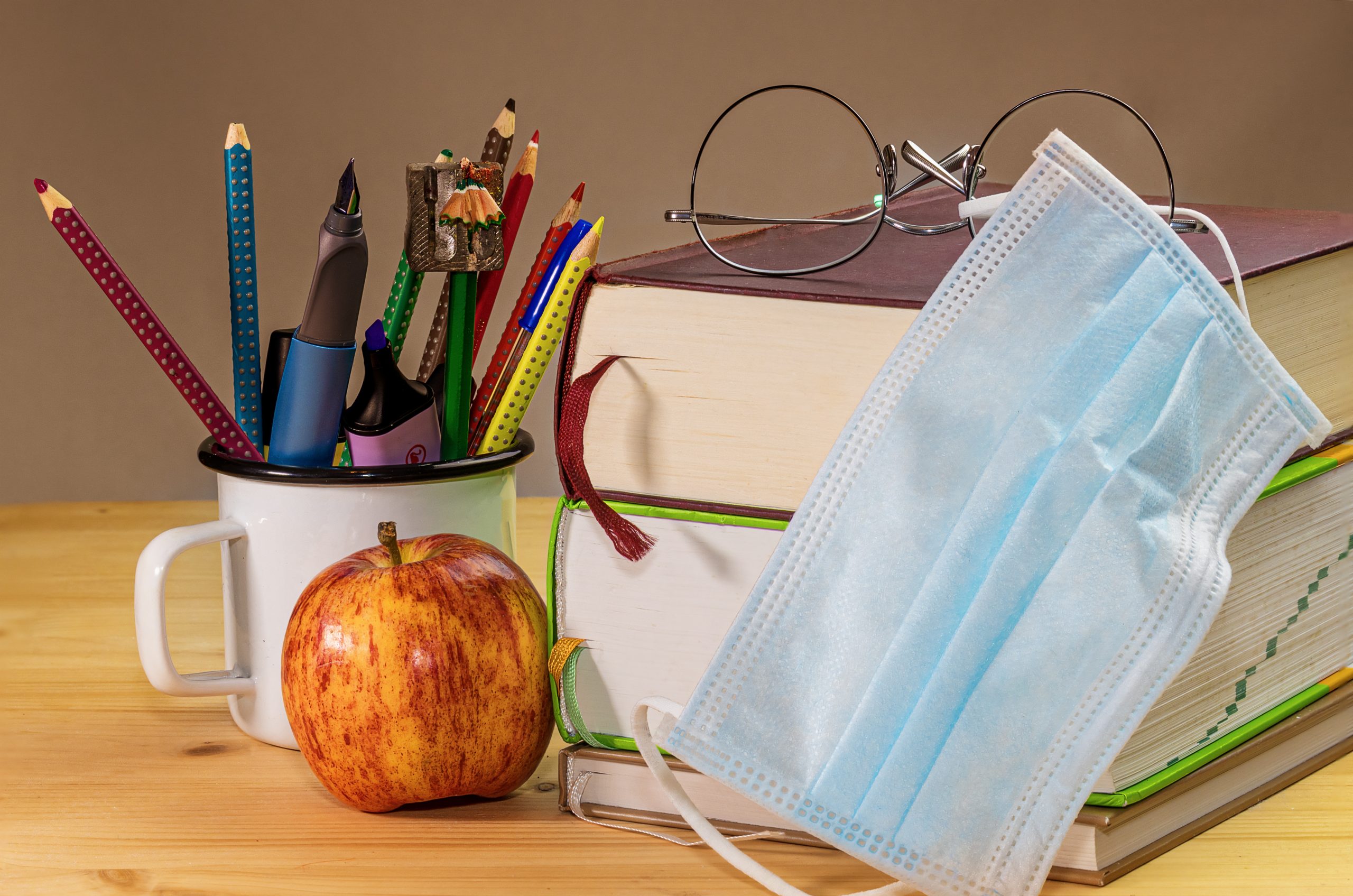 A desk with textbooks, a red apple, a cup full of pens and pencils, a pair of glasses, and a blue surgical mask gathered on top of it.