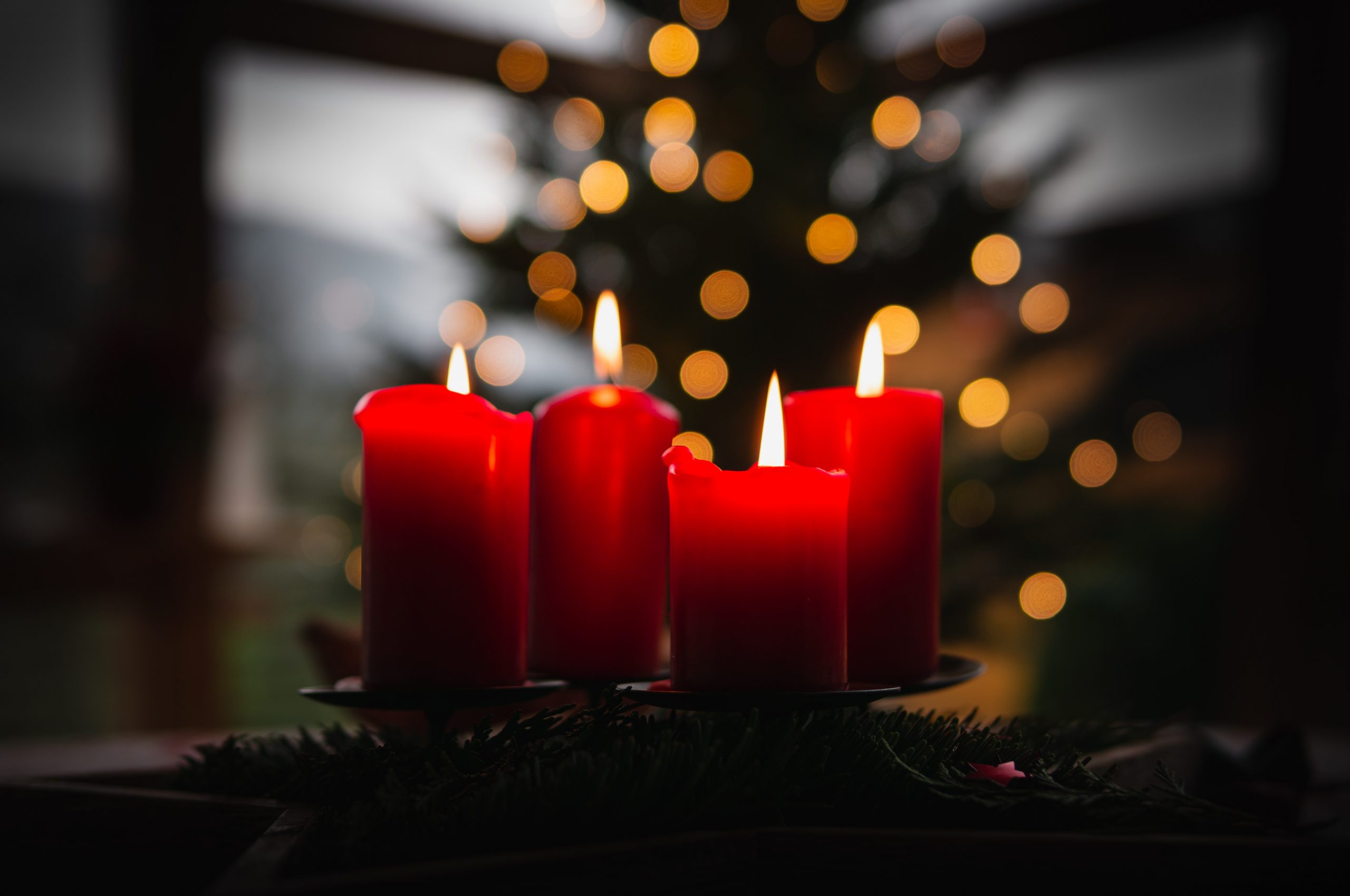 A cluster of festive red candles all glowing together in the dark, behind which a simple Christmas tree with golden string lights is visible.