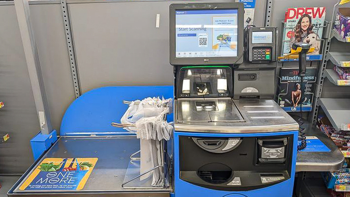 An empty blue and yellow self-checkout kiosk with a magazine rack on the right-hand side.