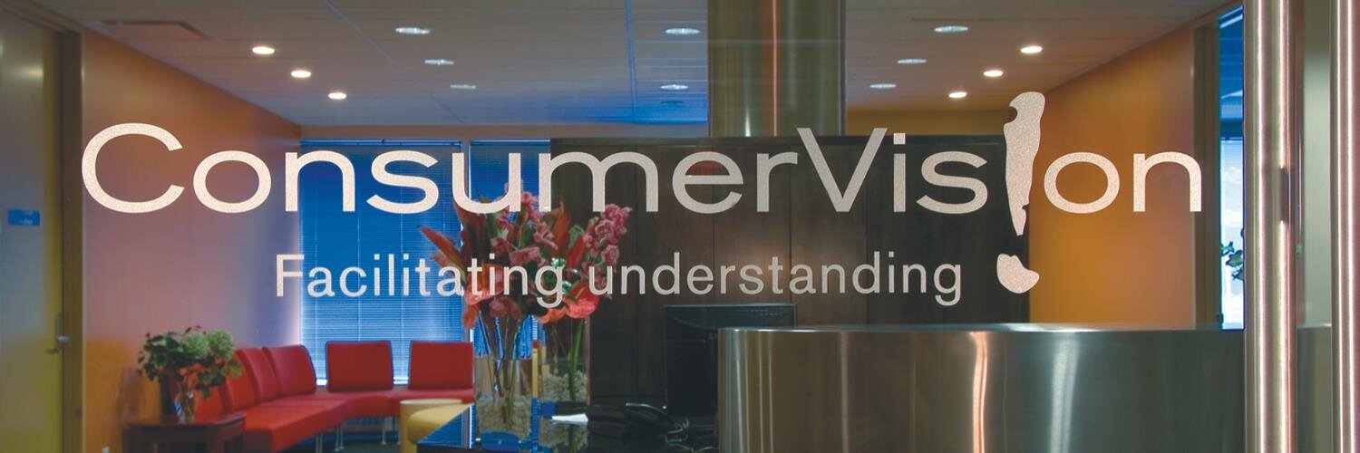 A photo of the front offices of Consumer Vision. They are behind a glass wall etched with the company name, where the second letter I in "Vision" has been changed to an exclamation mark, and displays their tagline: "Facilitating understanding."