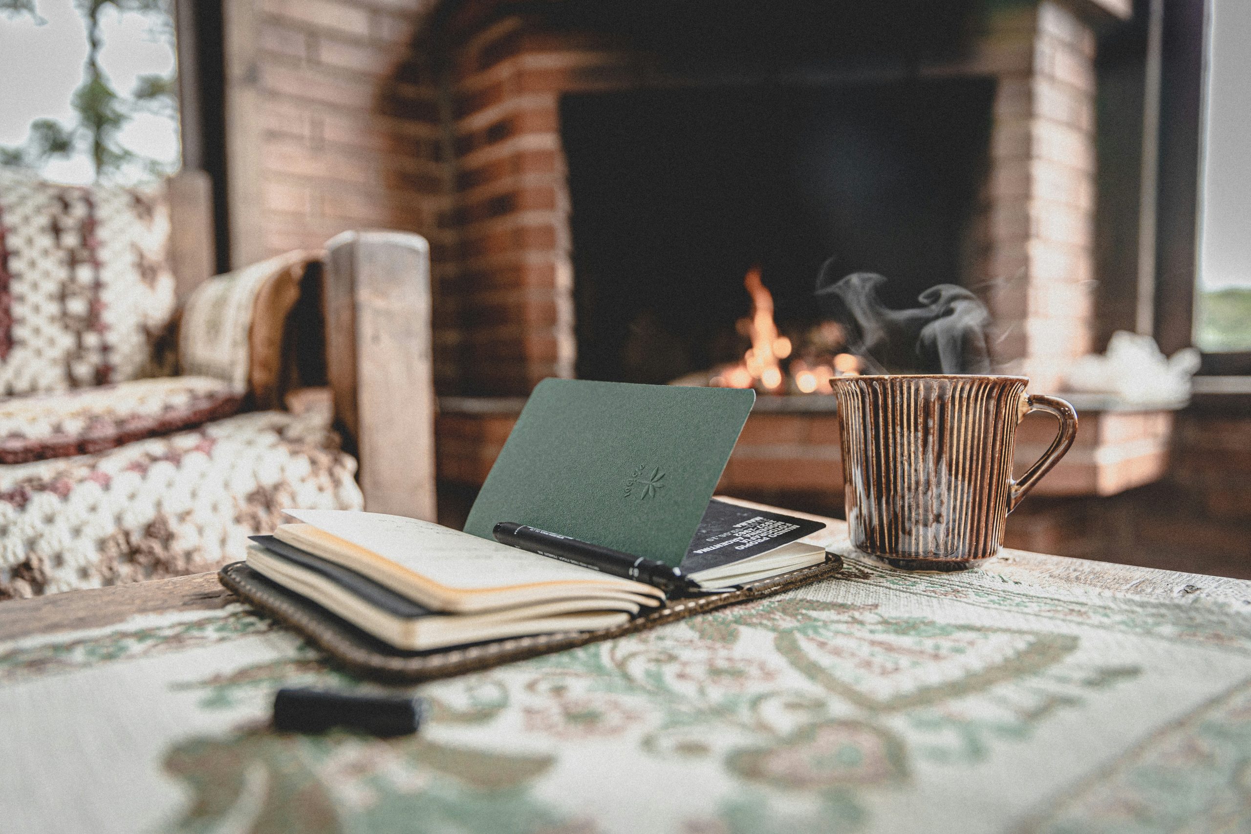 A view of a coffee table in a cozy living room. A cup of steaming tea sits next to an open journal, and a fireplace burns low in the background.
