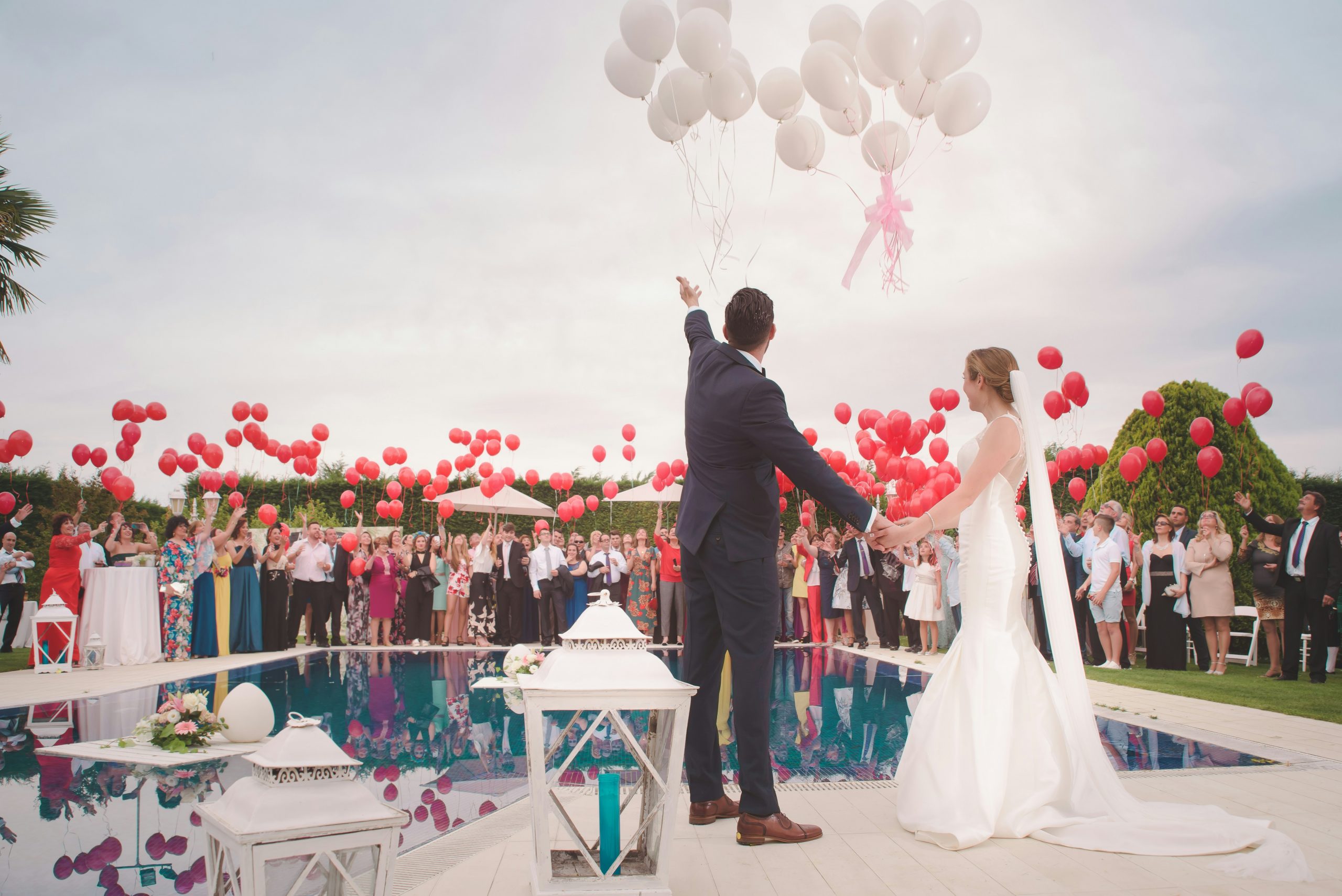 A bride and groom stand together in front of their assembled wedding guests. Everyone is joyfully releasing balloons into the sky.
