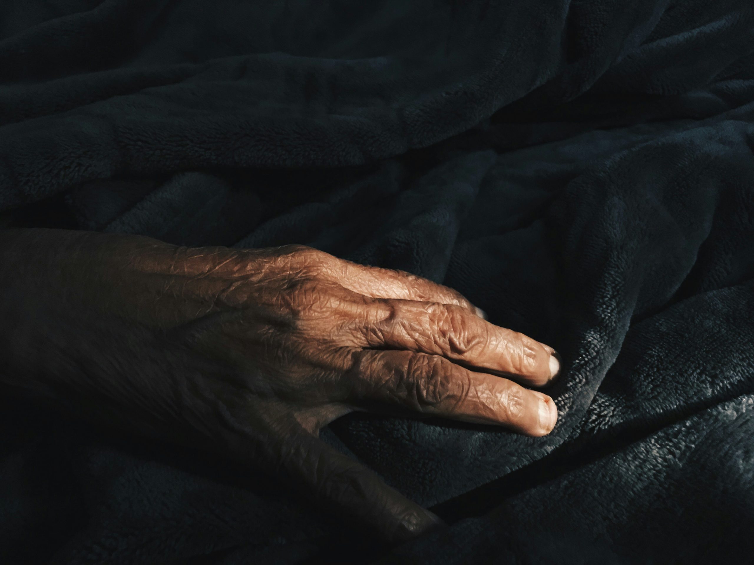 A close-up photo of an elderly person's hand reaching out from the dark.