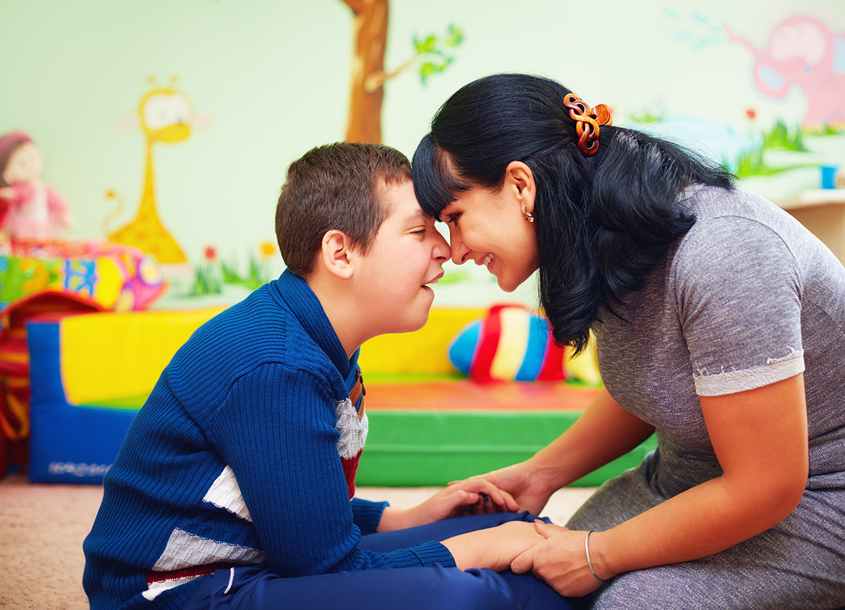 A young boy with developmental disabilities rests his forehead against that of a woman who holds his hands in hers.