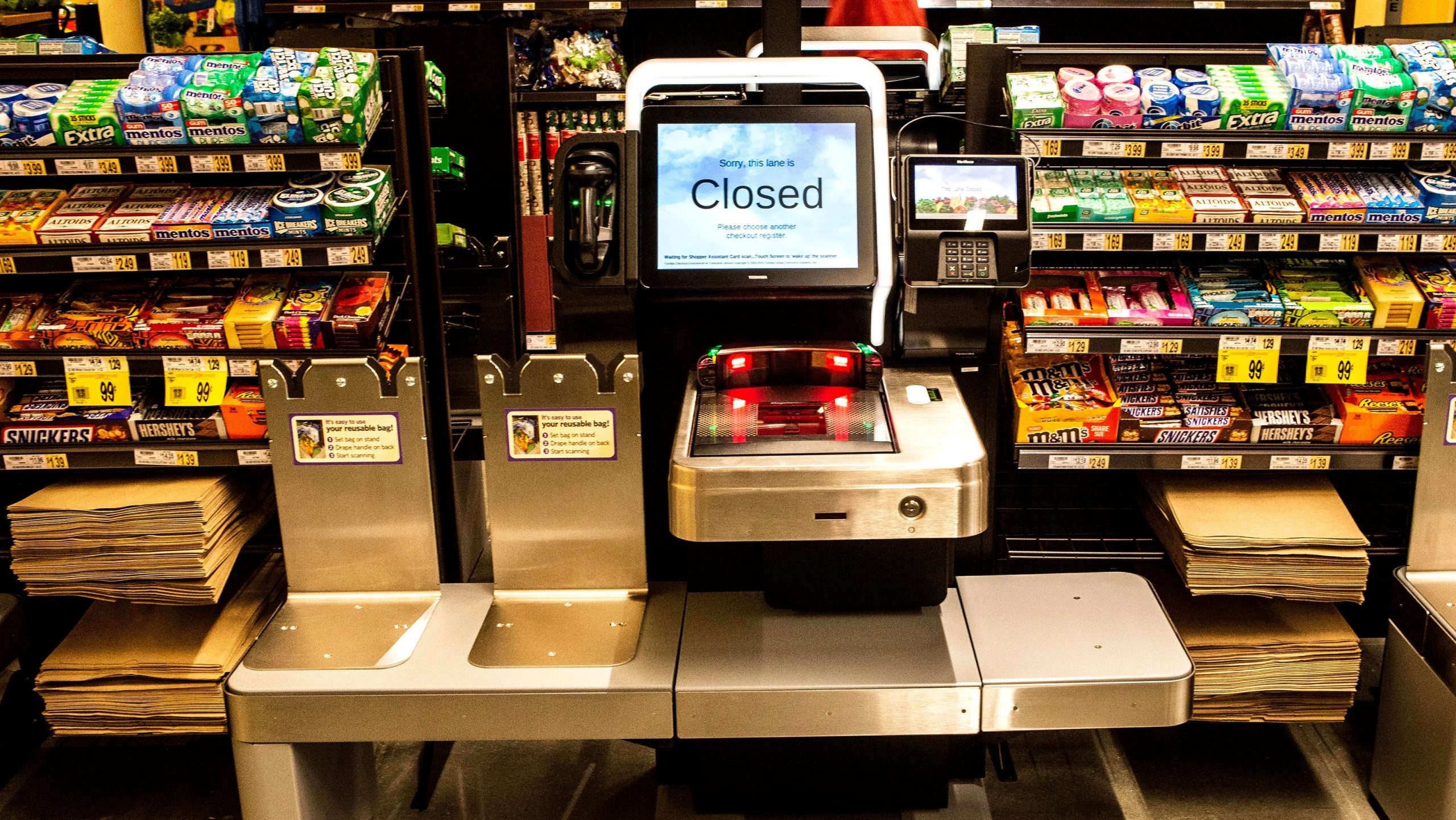 A self-checkout kiosk at a grocery store. The screen says, "Sorry, this is closed." There are colourful racks of chocolate bars and chewing gum behind the kiosk.