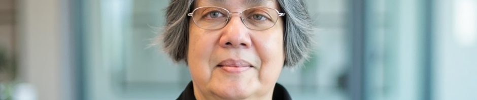 A photograph of Donna Jodhan with short hair and glasses pictured in a close-up portrait. She is wearing a black collared shirt and looking directly at the camera with a calm, neutral expression. The background is softly blurred, suggesting an indoor office setting with glass panels and modern furnishings, allowing the focus to remain on the subject.