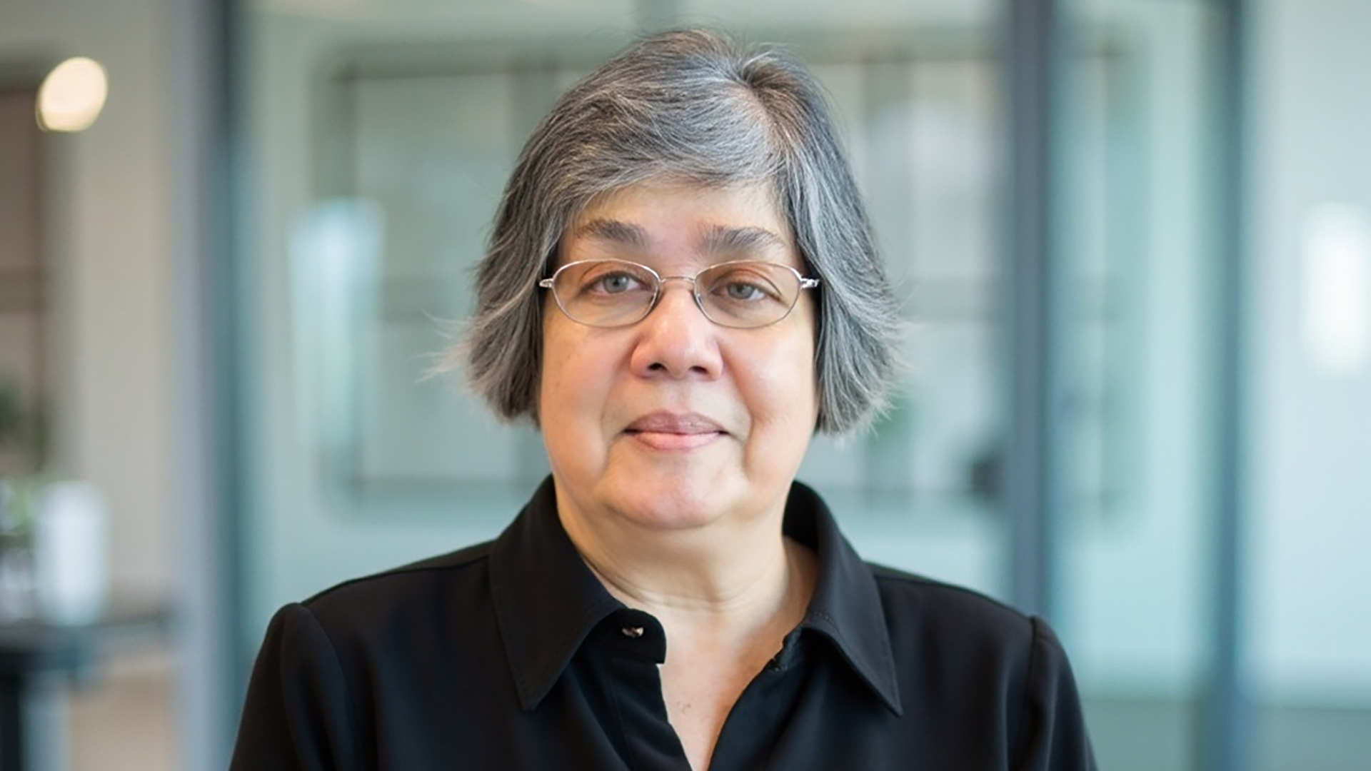 A photograph of Donna Jodhan with short hair and glasses pictured in a close-up portrait. She is wearing a black collared shirt and looking directly at the camera with a calm, neutral expression. The background is softly blurred, suggesting an indoor office setting with glass panels and modern furnishings, allowing the focus to remain on the subject.