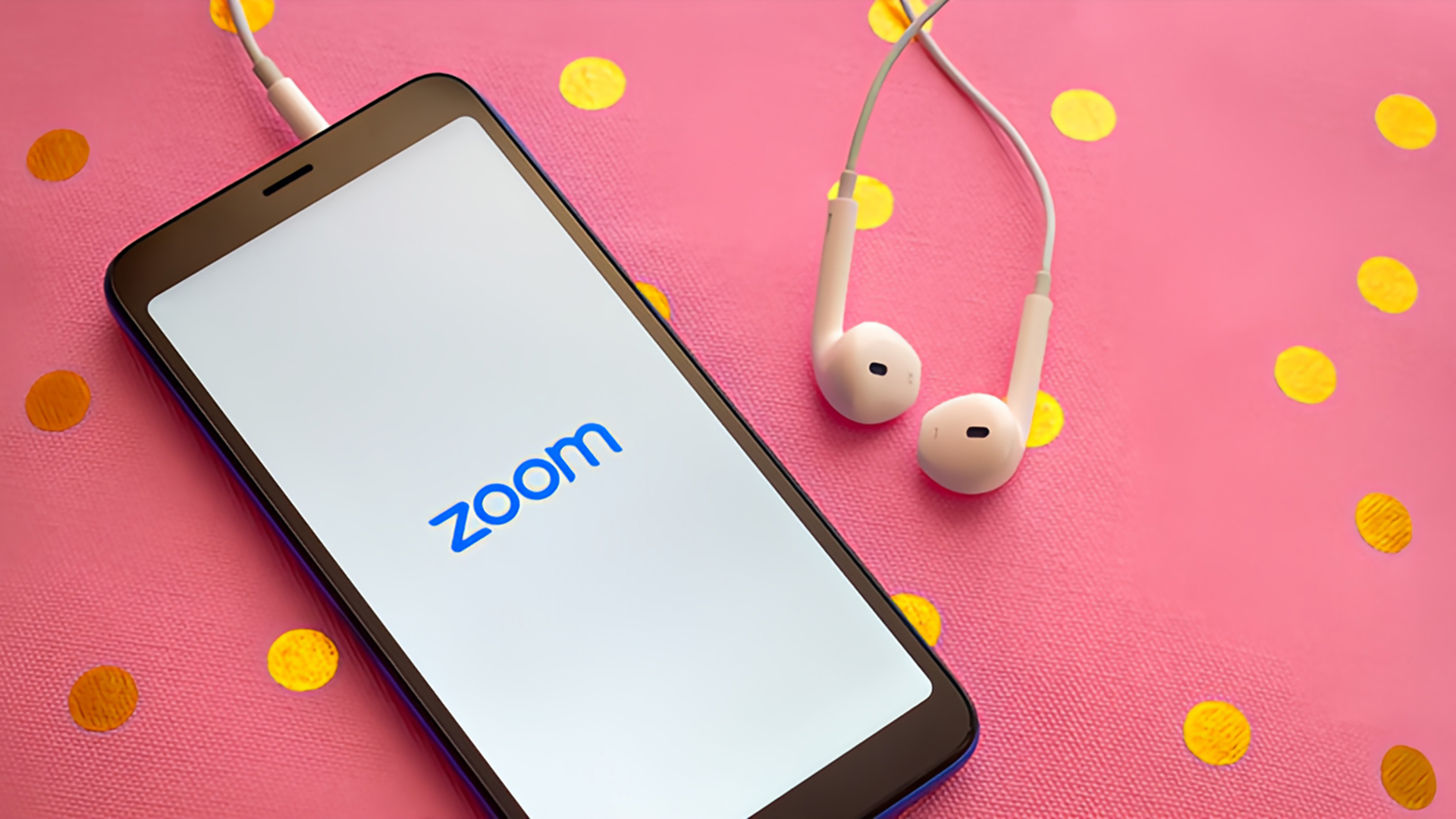 A smartphone with the Zoom app logo displayed on its screen lies on a pink surface decorated with scattered yellow polka dots. White wired earbuds are plugged into the phone and rest nearby, suggesting a setup for a virtual meeting or call. The bright and playful background contrasts with the clean, minimalist look of the phone screen, creating a casual yet tech-focused vibe.