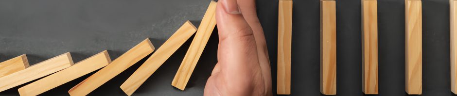A close-up image shows a hand positioned vertically between two rows of standing wooden dominoes on a dark surface, stopping the left row mid-fall. The dominoes on the left are already toppling, while the ones on the right remain upright, illustrating the concept of intervention or disruption in a chain reaction. The hand, emerging from a white shirt sleeve, symbolically represents control, prevention, or decision-making.