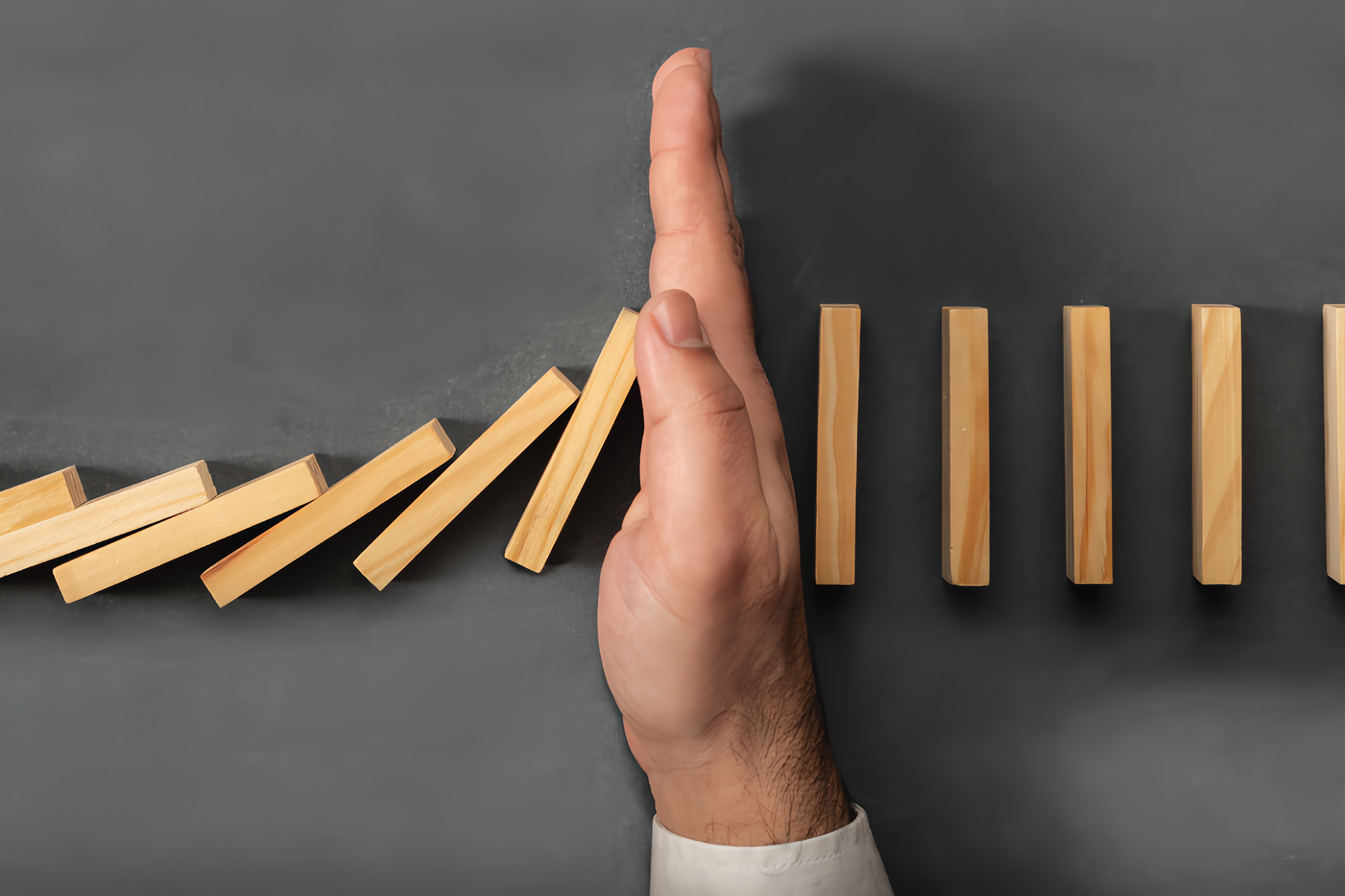 A close-up image shows a hand positioned vertically between two rows of standing wooden dominoes on a dark surface, stopping the left row mid-fall. The dominoes on the left are already toppling, while the ones on the right remain upright, illustrating the concept of intervention or disruption in a chain reaction. The hand, emerging from a white shirt sleeve, symbolically represents control, prevention, or decision-making.