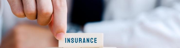 A close-up of a hand in a business suit stacking a wooden block labeled 'INSURANCE' on top of four other blocks. Each of the lower blocks features a blue icon representing different types of insurance: life (family icon), health (heart with a medical cross), home (house icon), and auto (car icon). The background is blurred, emphasizing the hand and blocks as the focal point.