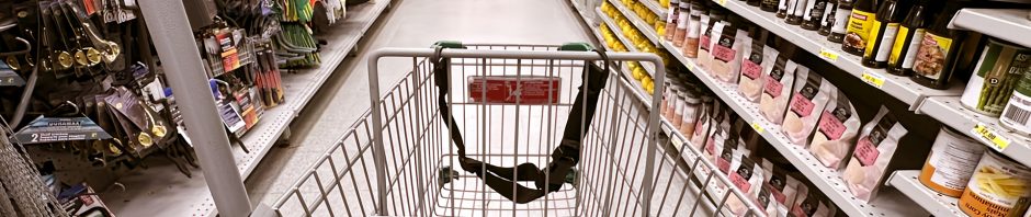 A shopping cart marked with the green and yellow logo of Dollarama is positioned in the center of a store aisle, with shelves on either side stocked with a variety of products. On the left, there are hardware and home improvement items such as gloves, tools, and hooks, while the right side features food items like sauces, canned goods, and packaged snacks. The aisle stretches ahead with bright lighting and a clean, polished floor, showcasing the store's well-organized layout.