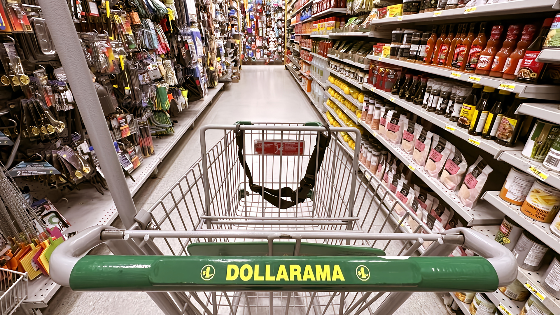 A shopping cart marked with the green and yellow logo of Dollarama is positioned in the center of a store aisle, with shelves on either side stocked with a variety of products. On the left, there are hardware and home improvement items such as gloves, tools, and hooks, while the right side features food items like sauces, canned goods, and packaged snacks. The aisle stretches ahead with bright lighting and a clean, polished floor, showcasing the store's well-organized layout.