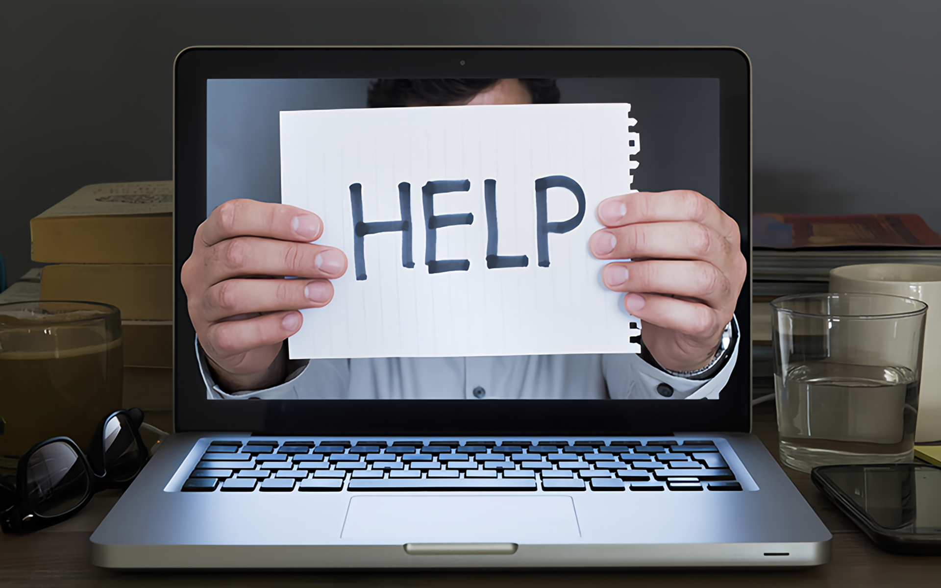 A laptop screen displays a man holding a white piece of lined paper with the word 'HELP' written in bold black letters, giving the illusion that his hands are emerging from the screen. The man’s face is partially obscured, and he is dressed in a white shirt. The laptop is placed on a desk cluttered with objects including a pair of glasses, a coffee mug, a glass of water, a smartphone, and stacked books, creating a scene that suggests stress, overwhelm, or a call for assistance in a work or academic setting.
