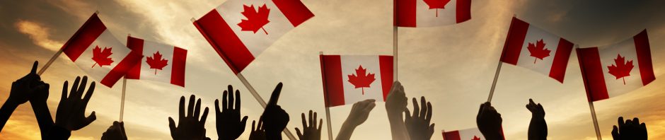 A crowd of people is seen from behind, raising their hands and waving Canadian flags against a dramatic sunset sky. The red and white flags, featuring the maple leaf emblem, stand out against the warm hues of orange and yellow in the background. Silhouettes of the individuals create a sense of unity and celebration, suggesting a patriotic gathering or national event. The positioning of the flags and the reaching hands add movement and energy to the scene.
