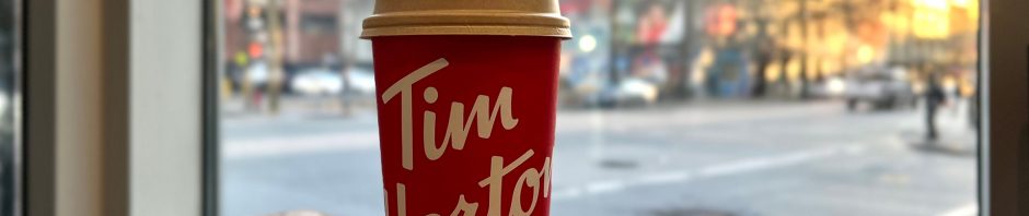 A red Tim Hortons coffee cup with a tan lid sits on a wooden table near a large window, with an urban street scene visible in the background; the cup features the brand's white cursive logo, and outside, a mix of blurry buildings, a green traffic light, and a few pedestrians and cars are seen in soft daylight, suggesting early morning or late afternoon.