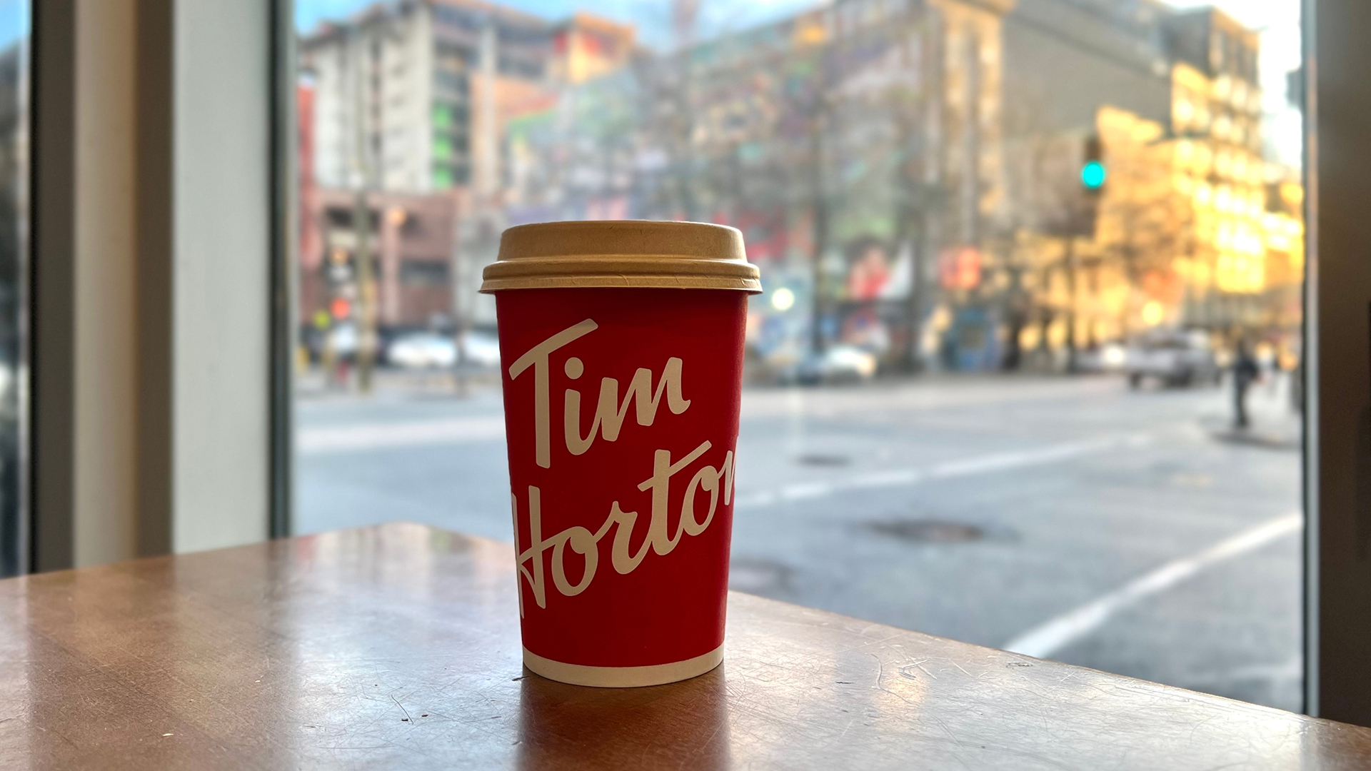 A red Tim Hortons coffee cup with a tan lid sits on a wooden table near a large window, with an urban street scene visible in the background; the cup features the brand's white cursive logo, and outside, a mix of blurry buildings, a green traffic light, and a few pedestrians and cars are seen in soft daylight, suggesting early morning or late afternoon.
