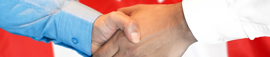 Two individuals are shaking hands firmly in front of a prominently displayed Canadian flag, symbolizing agreement, partnership, or diplomacy. One person wears a blue dress shirt while the other wears a white one, indicating a professional or formal context. The red and white flag with its bold red maple leaf in the center creates a strong national backdrop, emphasizing a Canadian connection or setting for the handshake.