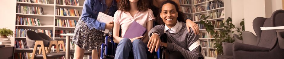 A group of five diverse young adults pose together in a cozy library or study room, with shelves of colorful books behind them; a woman in a light pink polo shirt and jeans sits in a wheelchair at the center, holding a purple notebook and smiling, while the others gather closely around her, smiling warmly and showing a sense of friendship and support.