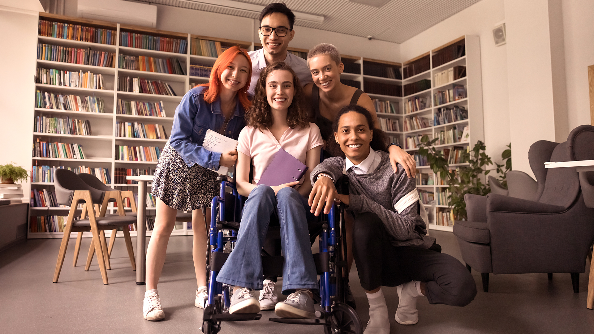 A group of five diverse young adults pose together in a cozy library or study room, with shelves of colorful books behind them; a woman in a light pink polo shirt and jeans sits in a wheelchair at the center, holding a purple notebook and smiling, while the others gather closely around her, smiling warmly and showing a sense of friendship and support.