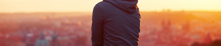 A person wearing a dark hoodie and jeans sits on a stone ledge, facing away from the camera and looking out over a blurred cityscape bathed in warm golden light from the setting sun; red and pink flowers frame the lower edge of the image, and the atmosphere suggests quiet reflection or solitude.