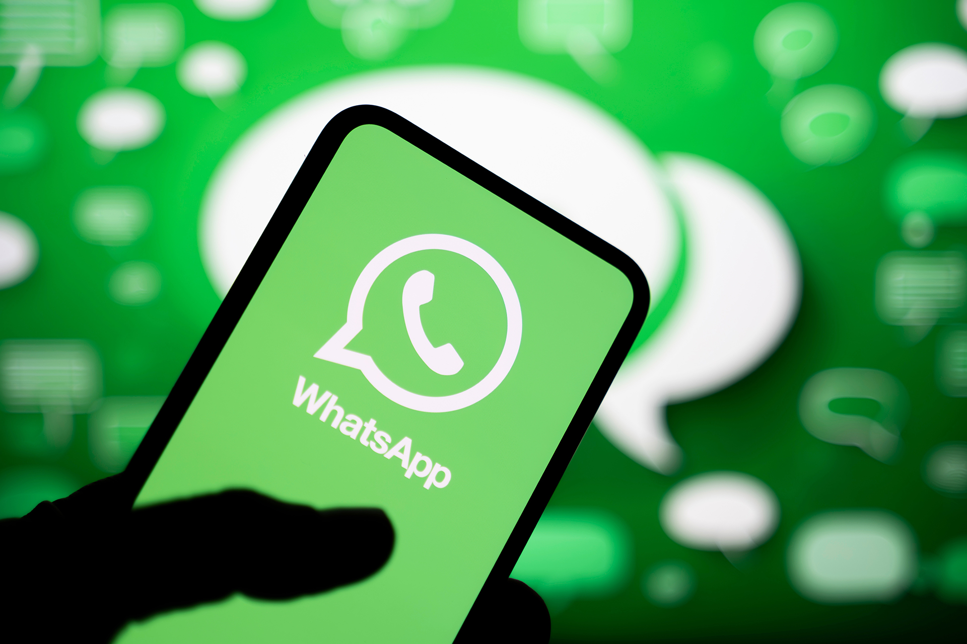 A silhouetted hand holds a tilted smartphone displaying the green WhatsApp screen with the white chat-bubble phone logo and the word WhatsApp, set against a bright green, softly blurred background filled with floating message icons and speech bubbles.