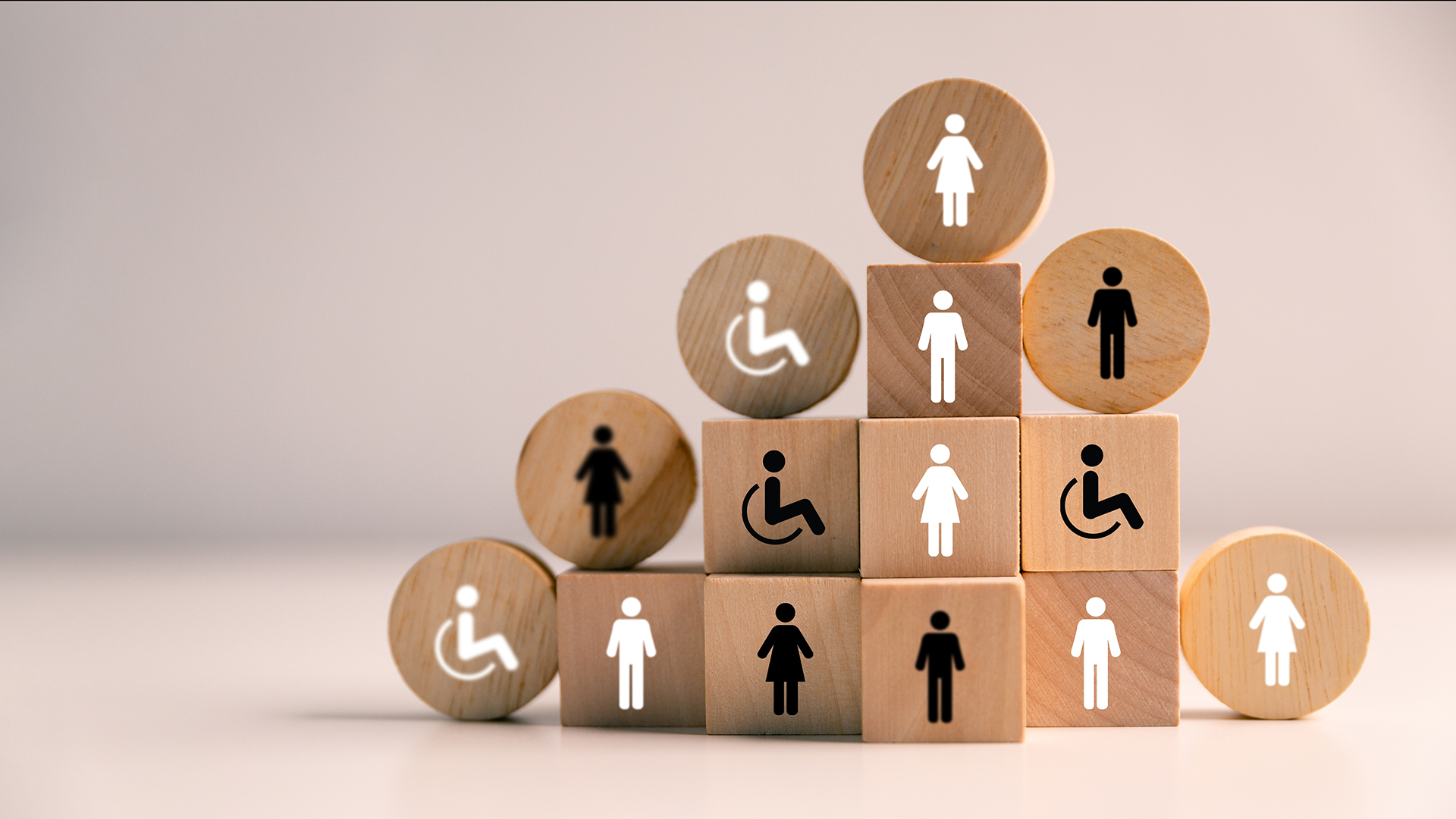 Stacked square and round wooden blocks arranged in a pyramid display simple black and white icons representing people, including male and female figures and disabled users, set against a soft neutral background, conveying themes of diversity, inclusion, and accessibility.