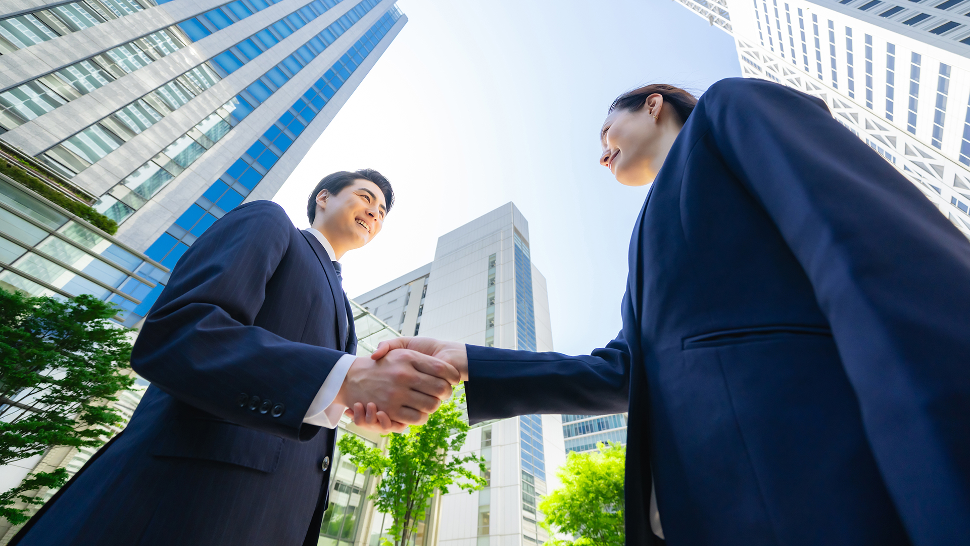 Two business professionals in dark suits shake hands outdoors in a modern city setting, surrounded by tall glass and steel buildings with green trees at their bases, under a bright blue sky.