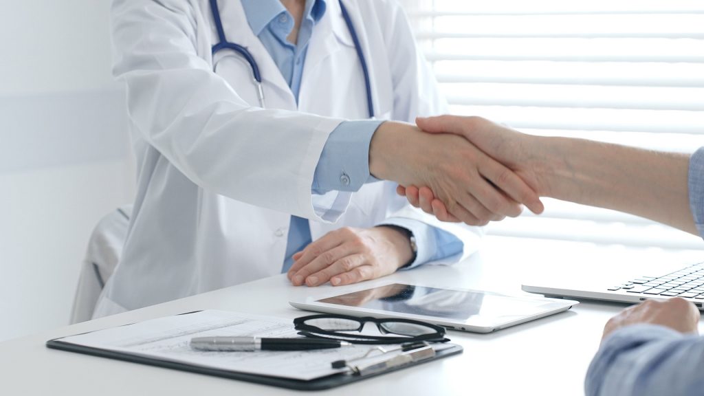 Doctor in a white coat with a stethoscope shakes hands with a patient across a bright clinic desk, suggesting a professional agreement or consultation, with a tablet, clipboard holding documents, pen, and eyeglasses in the foreground and a laptop to the side, while soft daylight filters through window blinds in the background.