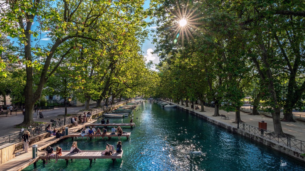 Tree-lined canal with clear blue-green water, small boats moored along the banks, and people relaxing on wooden docks and pathways under bright afternoon sun filtering through dense green leaves.