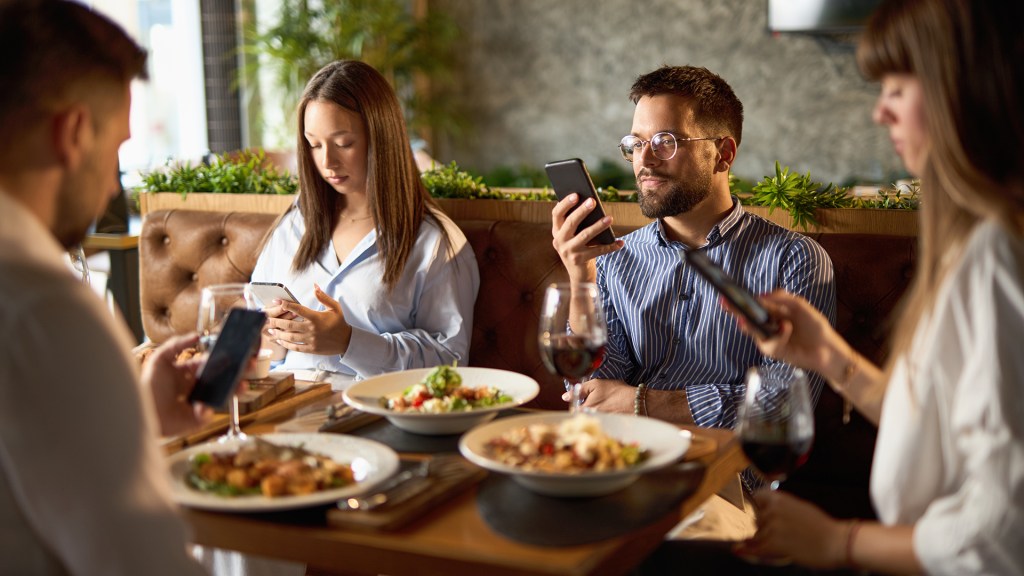 Four people sit around a restaurant table with plates of food and glasses of red wine, each person focused on their smartphone instead of interacting with one another, in a warmly lit modern dining setting with green plants in the background.