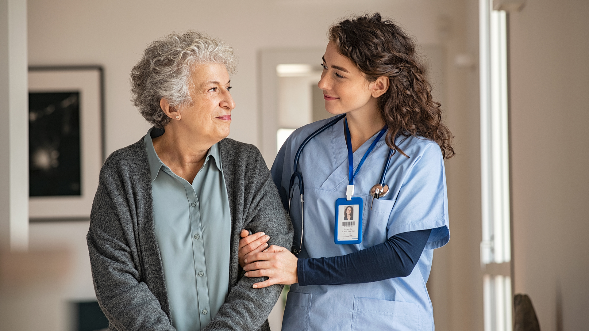 A smiling nurse in light blue scrubs with a stethoscope and ID badge gently holds the arm of an elderly woman with curly gray hair wearing a gray cardigan and light blue blouse, as they share a warm, caring look in a bright indoor setting.