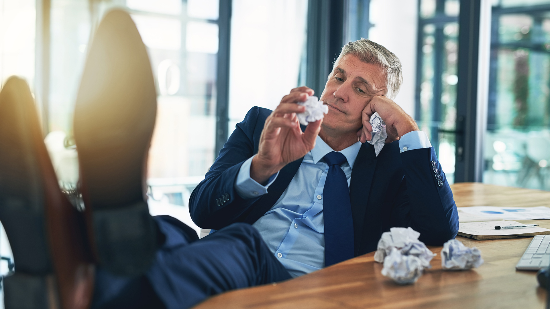 A middle-aged man in a blue suit sits at a desk in an office, looking bored and unmotivated with his feet propped up on the table. He holds a crumpled piece of paper in one hand and rests his head on the other, surrounded by several more crumpled papers on the desk alongside documents and a keyboard, with sunlight streaming through large windows in the background.