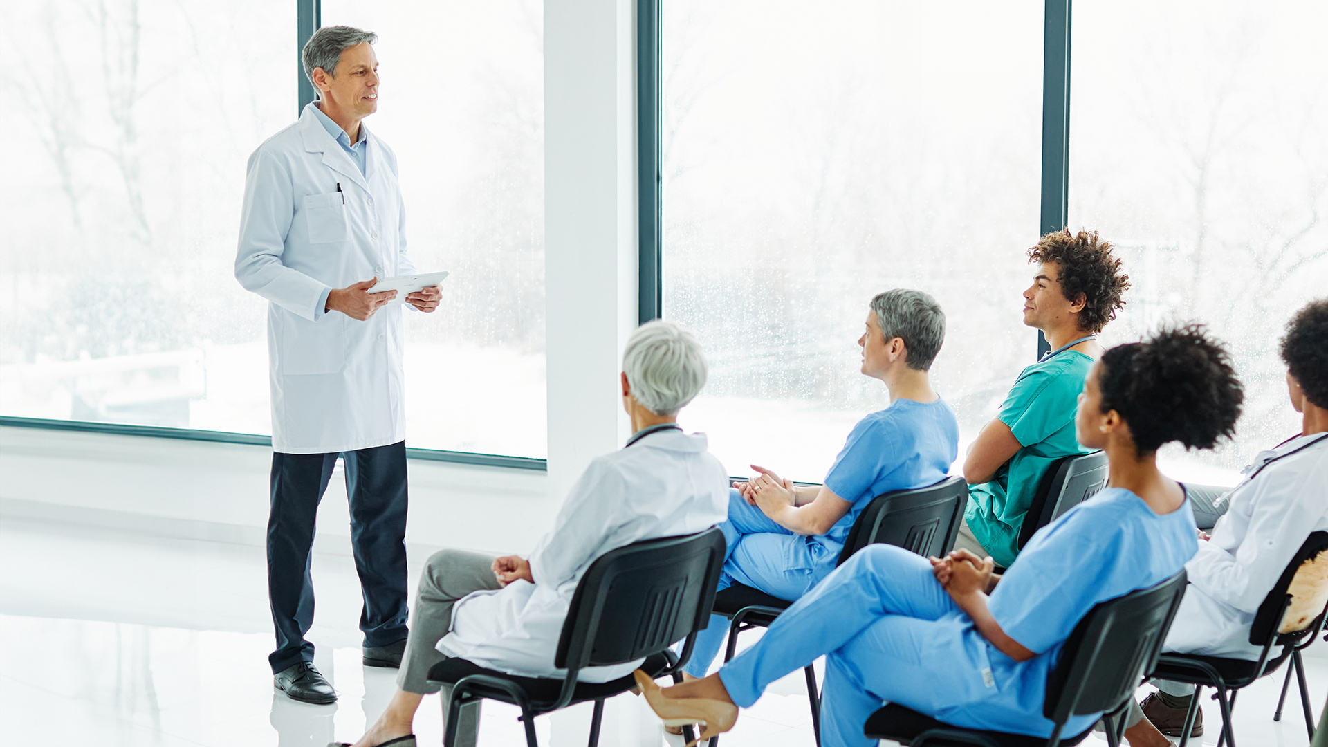 A male doctor in a white lab coat stands at the front of a bright, modern room holding a tablet and speaking to a small group of seated medical staff in scrubs, including men and women of varied ages and ethnicities, who listen attentively in black chairs, with large frosted windows behind them diffusing soft daylight and creating a clean, clinical atmosphere.