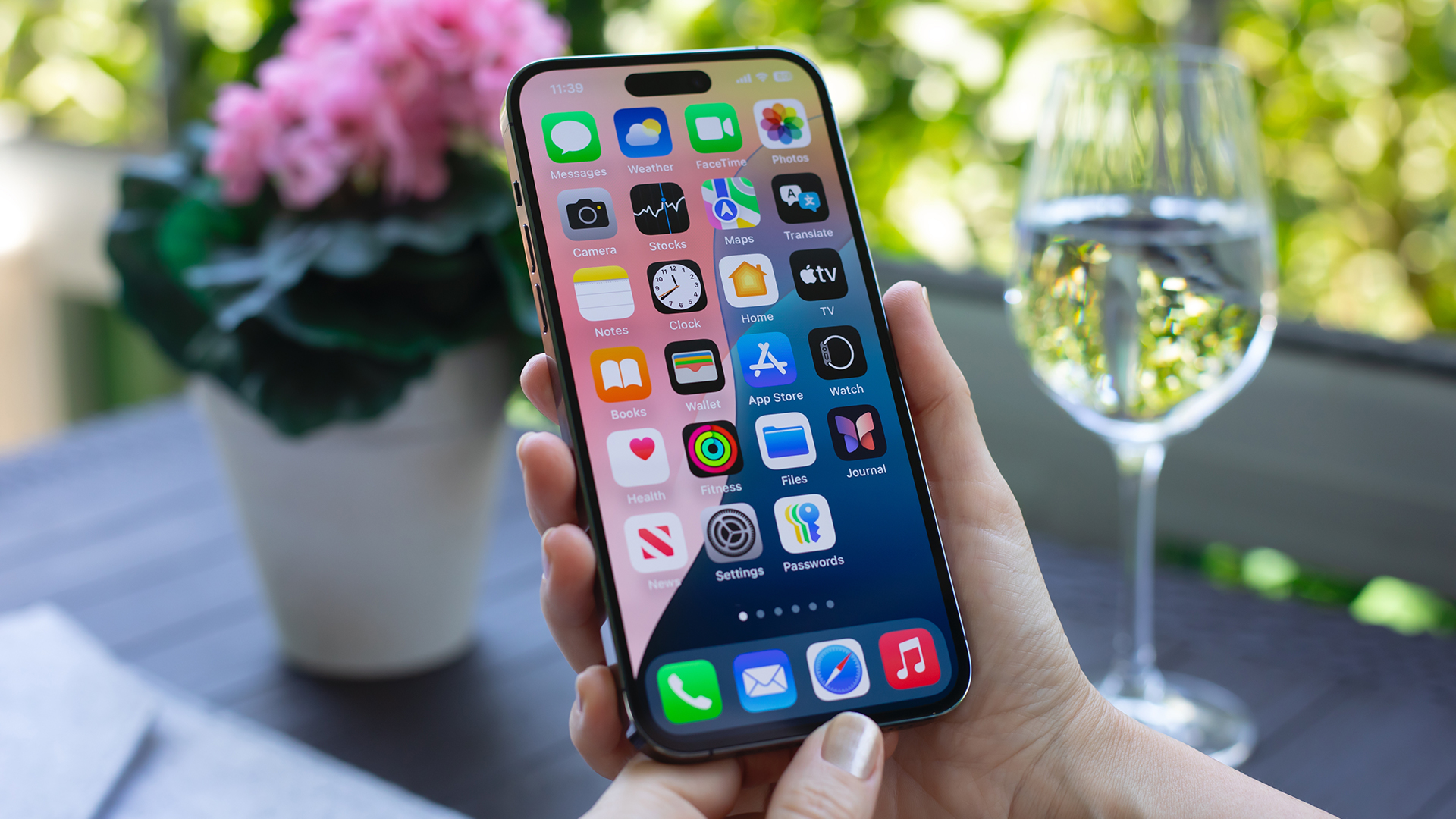 A person holds a modern smartphone displaying a grid of app icons on its home screen while seated outdoors at a table, with a blurred potted plant featuring pink flowers on the left and a clear wine glass filled with water on the right, set against a bright green garden background in natural daylight.